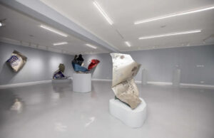 Gallery with abstract metallic sculptures on pedestals, featuring crumpled forms in silver, blue, and red. Bright lighting and minimalistic decor set a modern tone.