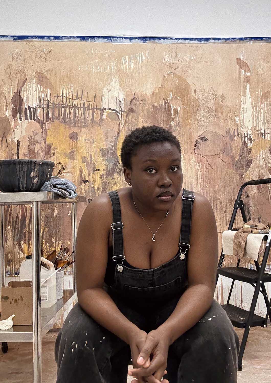 A person in paint-splattered overalls sits pensively in front of a large abstract painting. The setting is a creative studio, conveying focus and introspection.