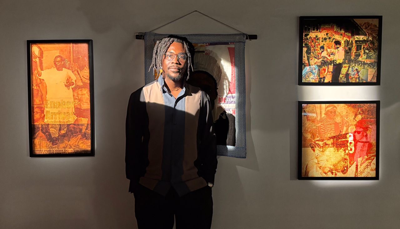 Alt text: Man with dreadlocks and glasses stands in dim gallery in front of a hanging artworks. Three colourful, abstract artworks are displayed on the wall, illuminated by overhead lighting.