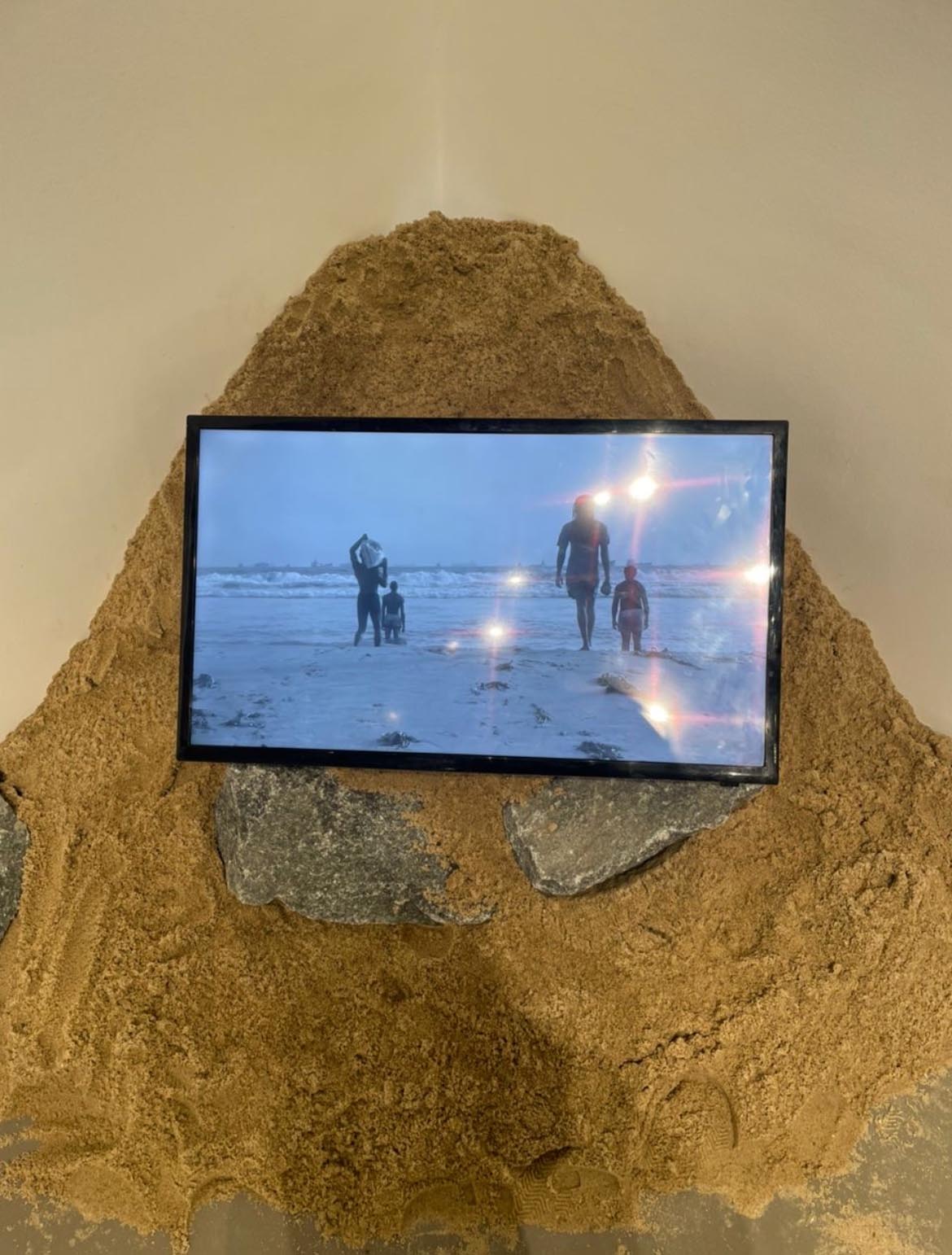 A TV screen embedded in a sand and rock mound shows a silhouette of a family walking on a beach. Glare on the screen adds a dreamy effect.